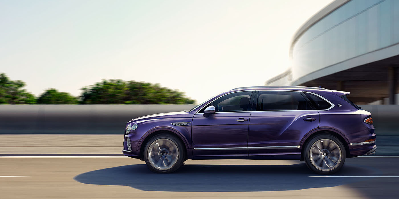 Bentley Monterrey Bentley Bentayga Extended Wheelbase Mulliner luxury SUV profile in Tanzanite Purple paint, driving on an country road past trees.